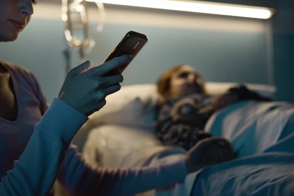 年轻女子晚上在医院帮助她的孩子 她正在和她的智能手机聊天 并在网上联系 — 图库照片