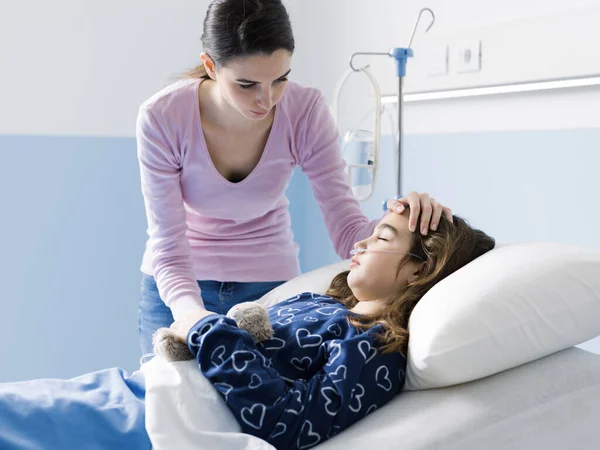 Young woman comforting her sick child lying in bed at the hospital, she is touching her head, pediatrics and healthcare concept