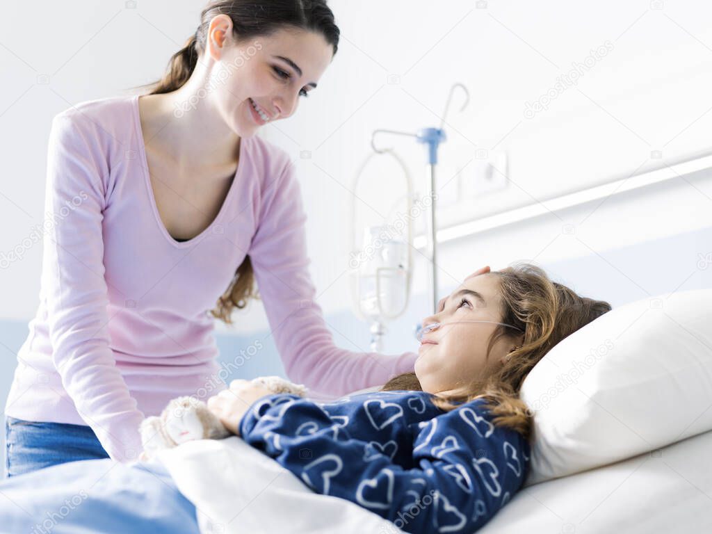 Young woman assisting her sick child lying in bed at the hospital, pediatrics and healthcare concept
