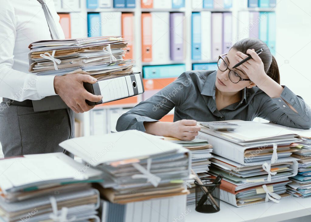 Young stressed secretary in the office overwhelmed by work and desk full of files, her boss is bringing more paperwork to her