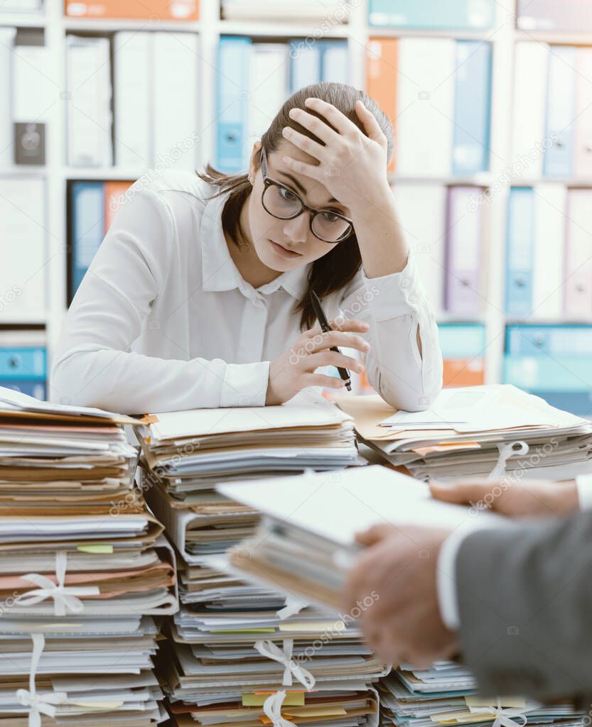 Exhausted young secretary overloaded with work, her boss is bringing more paperwork to her