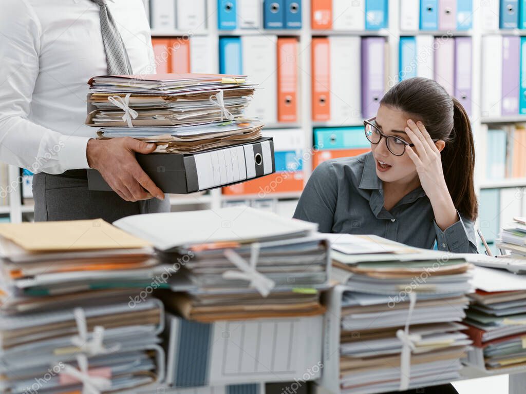 Young stressed secretary in the office overwhelmed by work and desk full of files, her boss is bringing more paperwork to her