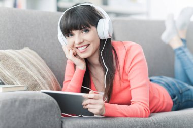 Kanepede dinlenen gülümseyen genç bir kadın, kulaklık takıyor, dijital tablet kullanıyor ve internetten film izliyor.