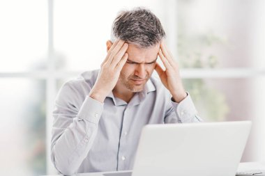 Ofis masasında çalışan stresli iş adamı baş ağrısı çekiyor, şakaklarına dokunuyor.