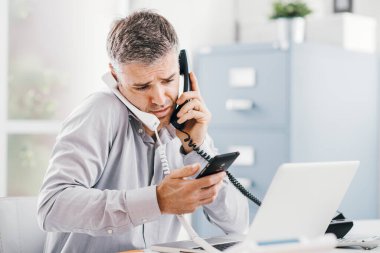 Stressed desperate businessman working in his office and having multiple calls, he is holding two handsets and a mobile phone, business management concept clipart