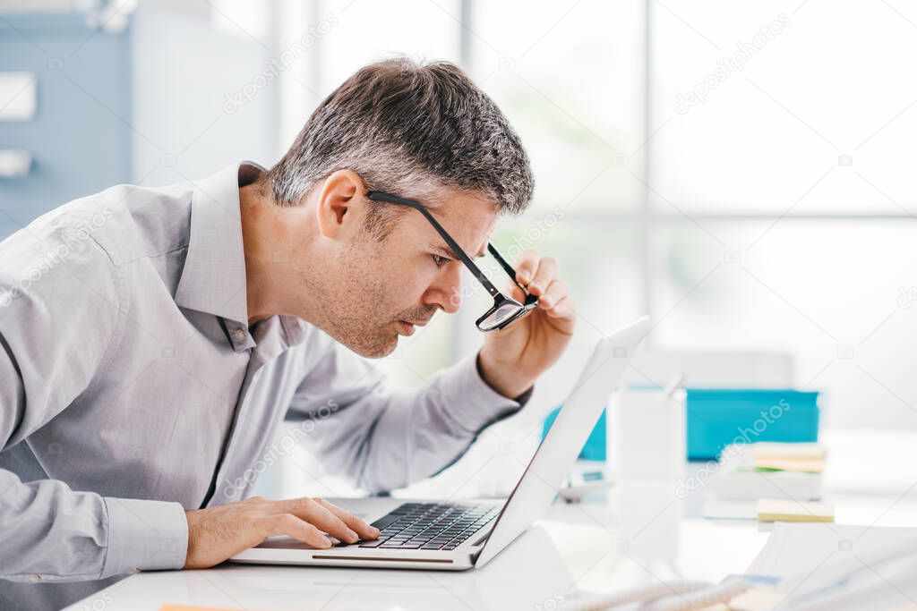 Businessman working at office desk, he is staring at the laptop screen close up and holding his glasses, workplace vision problems