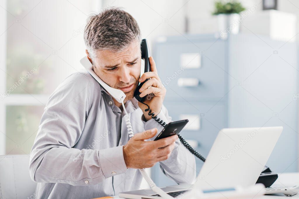 Stressed desperate businessman working in his office and having multiple calls, he is holding two handsets and a mobile phone, business management concept