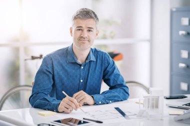 Profesyonel mimar ofis masasında oturuyor ve çalışıyor. Bir proje planı, mühendislik ve mimari konseptini kontrol ediyor.