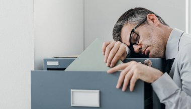 Tired lazy office worker leaning on a filing cabinet and sleeping, he is falling asleep standing up; stress, unproductivity and sleep disorders concept clipart