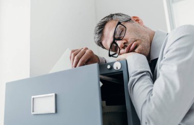 Tembel bir ofis çalışanı dosya dolabına yaslanıp uyuyor, ayakta uyuyor; stres, verimsizlik ve uyku bozuklukları kavramı