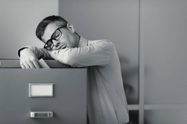 Tired lazy office worker leaning on a filing cabinet and sleeping, he is falling asleep standing up; stress, unproductivity and sleep disorders concept clipart