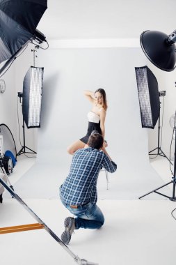 Stüdyoda profesyonel fotoğraf çekimi: güzel genç bir model gülümsüyor ve poz veriyor; fotoğrafçı dijital bir kamerayla fotoğraf çekiyor