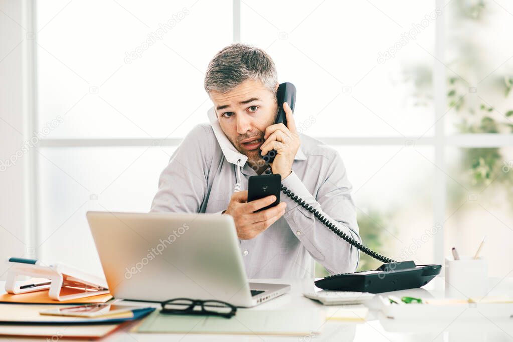 Stressed desperate businessman working in his office and having multiple calls, he is holding two handsets and a mobile phone, business management concept