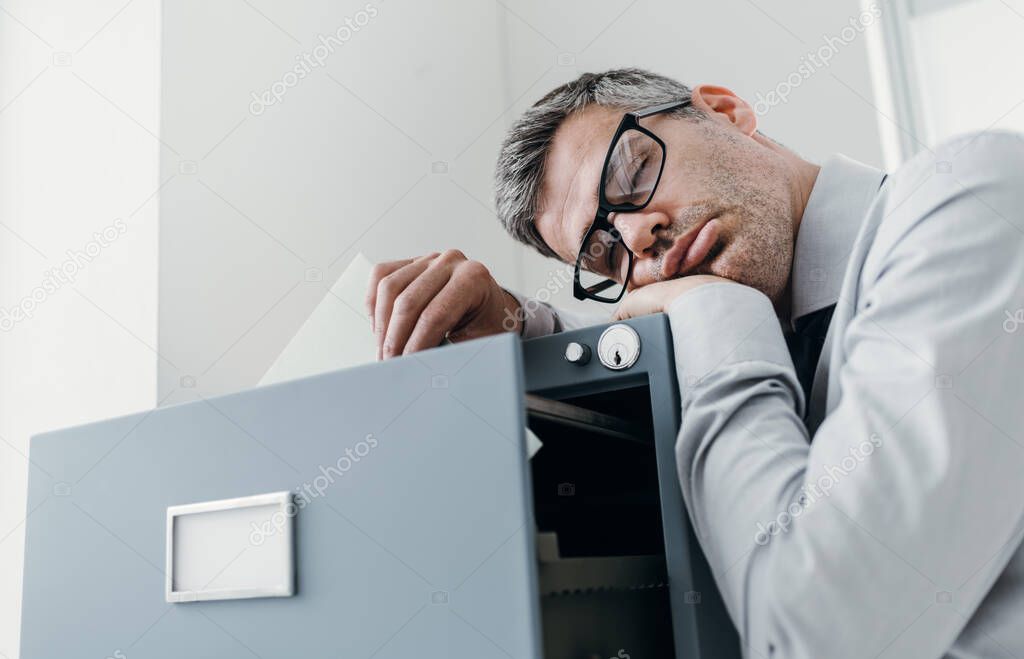Tired lazy office worker leaning on a filing cabinet and sleeping, he is falling asleep standing up; stress, unproductivity and sleep disorders concept