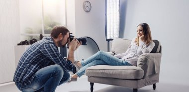 Stüdyoda profesyonel fotoğraf çekimi, bir kadın model koltukta poz veriyor ve gülümsüyor; fotoğrafçı dijital kamerasıyla fotoğraf çekiyor.