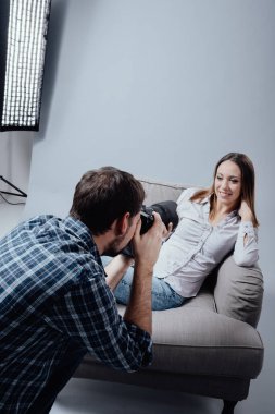 Stüdyoda profesyonel fotoğraf çekimi, bir kadın model koltukta poz veriyor ve gülümsüyor; fotoğrafçı dijital kamerasıyla fotoğraf çekiyor.