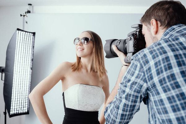 Профессиональная фотосессия в студии: красивая молодая модель позирует в солнечных очках и улыбается, фотограф снимает фотоаппаратом
