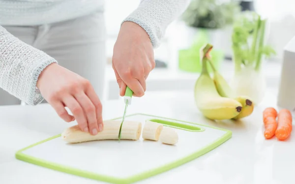 彼女の台所で健康的なビーガンスナックを準備女性 彼女はナイフでまな板にバナナをスライスしています — ストック写真