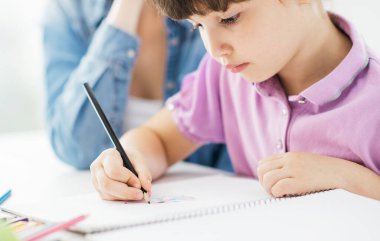 Anne ve çocuk evde birlikte vakit geçiriyorlar, kız kalemle bir defter çiziyor.