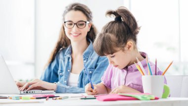 Mutlu anne ve çocuk evde birlikte vakit geçiriyor, kadın dizüstü bilgisayarla çalışıyor ve kız resim çiziyor.