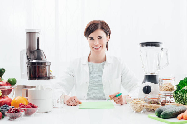 Smiling professional nutritionist holding an apple, she has healthy fruits, vegetables and juicers on her desk; healthy diet and wellness concept