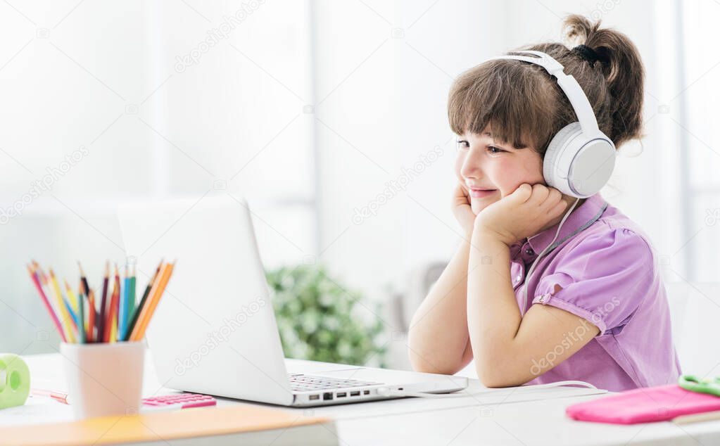 Happy cute girl watching videos streaming online on her laptop at home, she is smiling and wearing headphones