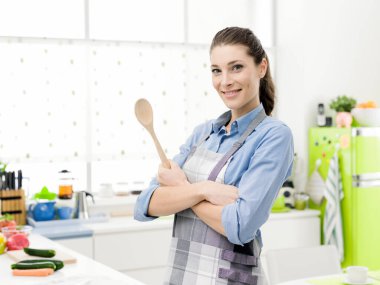 Kendine güvenen, önlüklü genç bir kadın mutfakta poz veriyor, yemek hazırlıyor ve bir kaşık tutuyor.