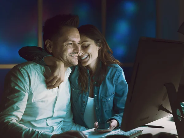 Happy romantic couple at home, they are connecting with their computer and cuddling, they are laughing together