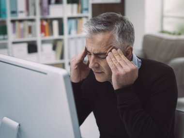 Yorgun iş adamı kötü bir baş ağrısı çekiyor, şakaklarına masaj yapıyor: stres ve teslim tarihi kavramı