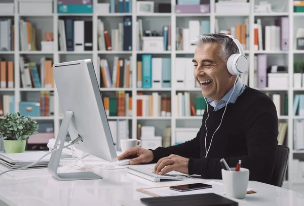 幸せな男はヘッドフォンを着用し 自宅のオフィスでオンラインで映画を見て 彼は楽しんで笑っています — ストック写真