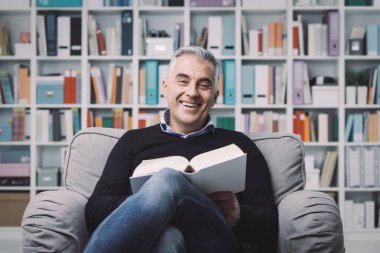 Gülümseyen kendine güvenen bir adam koltukta rahatlıyor ve bir kitap okuyor, arka planda kitaplıklar var.