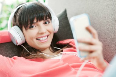Evdeki kanepede oturan çekici genç kadın, akıllı telefonuyla müzik çalıyor ve kulaklık takıyor, boş zaman ve eğlence konsepti kullanıyor.