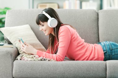 Kulaklıklı genç bir kadın kanepede dinleniyor ve internette akıllı telefon kullanarak müzik dinliyor.
