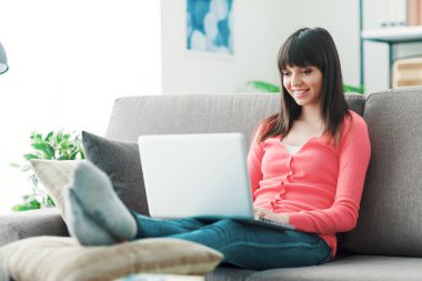 Genç ve çekici bir kadın evde dinleniyor, kanepede oturuyor ve bir dizüstü bilgisayar kullanarak internete bağlanıyor