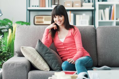 Evde gülümseyen çekici bir kadın, kanepede oturuyor, rahatlıyor ve poz veriyor.