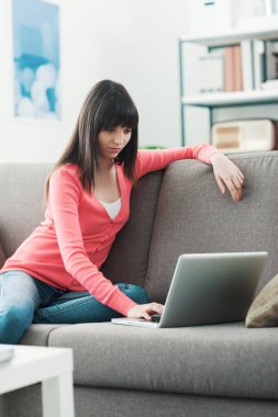 Genç ve çekici bir kadın evde dinleniyor, kanepede oturuyor ve bir dizüstü bilgisayar kullanarak internete bağlanıyor