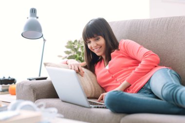 Evde gülümseyen genç bir kadın, kanepede rahatlıyor ve bir dizüstü bilgisayarla sosyal iletişim kuruyor.