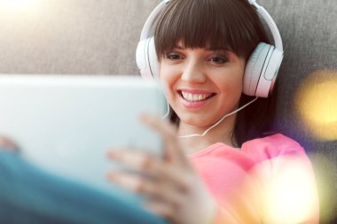 Evde kanepede dinlenen gülümseyen genç kadın, kulaklık takıyor, dijital tablet kullanıyor, video izliyor ve çevrimiçi müzik dinliyor.