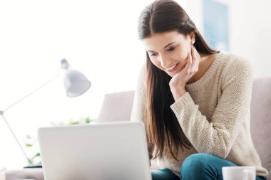 Oturma odasında gülümseyen genç bir kadın, kanepede oturuyor ve dizüstü bilgisayar kullanıyor.