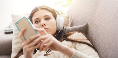Çekici kız kanepede dinleniyor ve akıllı telefonunda uygulamalar kullanıyor. Kulaklık takıyor.