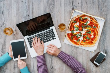 Ofis çalışanları ofiste yemek molası veriyorlar, pizza yiyorlar, dizüstü bilgisayar ve dijital tablet kullanıyorlar, düz konum alıyorlar.
