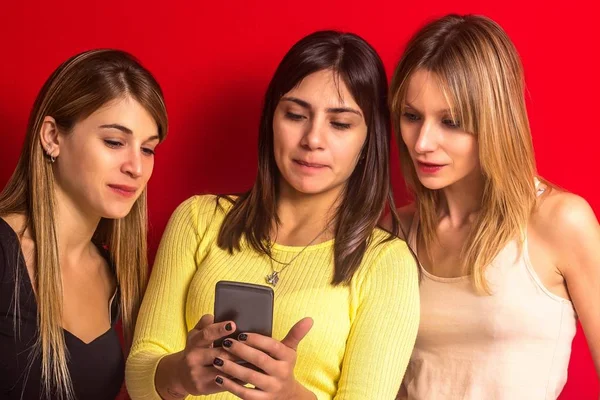 Tres chicas mirando un teléfono inteligente frente a la pared roja — Foto de Stock