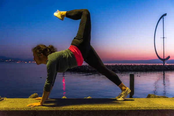Sarışın kadın İsviçre 'de Lozan' da gün batımında bir gölün önünde yoga egzersizleriyle sırtını geriyor.