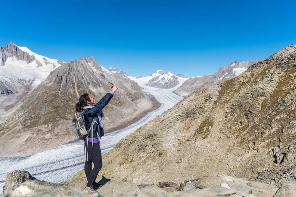 Sırtında sırt çantasıyla genç beyaz bir kadın arkasında devasa Aletsch Buzulu ile selfie çekiyor..