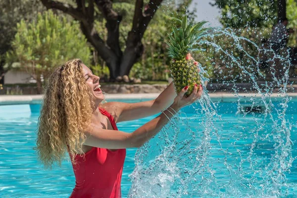 一个金发碧眼的女孩穿着红色泳衣在游泳池里从水里提起菠萝 形成了一条水滴的小径 — 图库照片