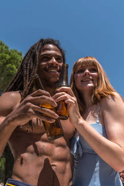 一个年轻女子与一个留着可怕头发的非洲男人坐在一起 在室外游泳池里举着一瓶啤酒敬酒的纵向照片 — 图库照片