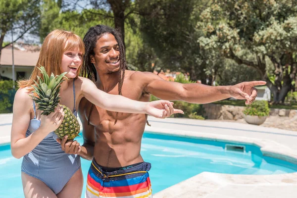 一个红头发的女孩和一个害怕的非洲男孩站在室外游泳池里 手里拿着一个菠萝 指着前面 — 图库照片