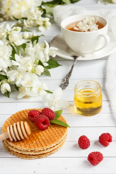 Tazza da tè con fiori di gelsomino e cialde sottili con miele e lampone su uno sfondo di legno chiaro — Foto Stock