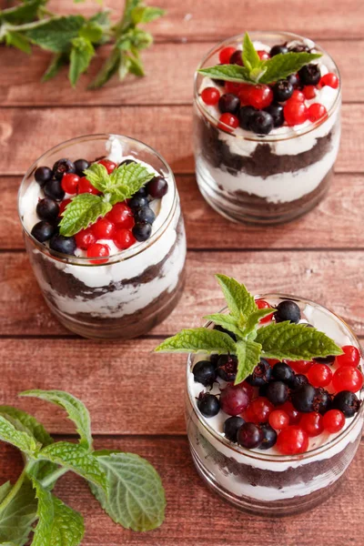 A sweet dessert of sponge cake with cream in a glass with fresh berries on wooden dark background