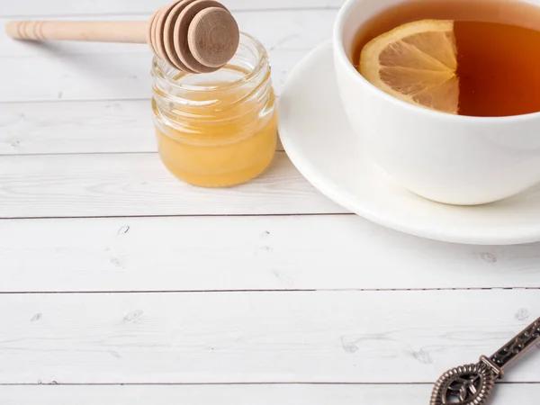Tè nero alle erbe in una tazza bianca, miele in un barattolo di limone fresco su sfondo di legno bianco — Foto Stock